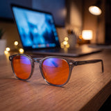 Aura blue light glasses resting on a wooden desk at night in front of a laptop, orange lenses showing their violet anti-reflective sheen