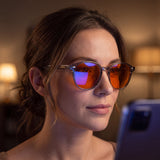 Woman wearing Aura blue light glasses while reading her phone at night, violet blue-light-blocking sheen visible on the orange lenses