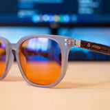 Close-up of Ash frosted grey square blue light glasses on a desk, orange lenses showing the violet coating sheen in front of a monitor