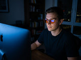 Man working late at a bright iMac in orange-lens blue light glasses, the screen reflected in the lenses