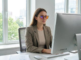 Woman at an office desktop wearing orange-lens blue light glasses, daylight from the window behind her