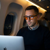 Traveller working on a laptop on a night flight, wearing Ember blue light glasses with orange lenses
