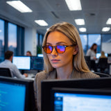 Woman at an open-plan office desk in Hazel tortoise round blue light glasses, orange lenses reflecting the monitors