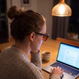 Woman typing at a laptop in the evening wearing the Rowan blue light glasses — glossy black panto TR90 frame