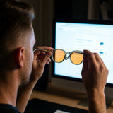 Man holding up Vesper blue light glasses in front of a bright monitor, orange lenses catching light